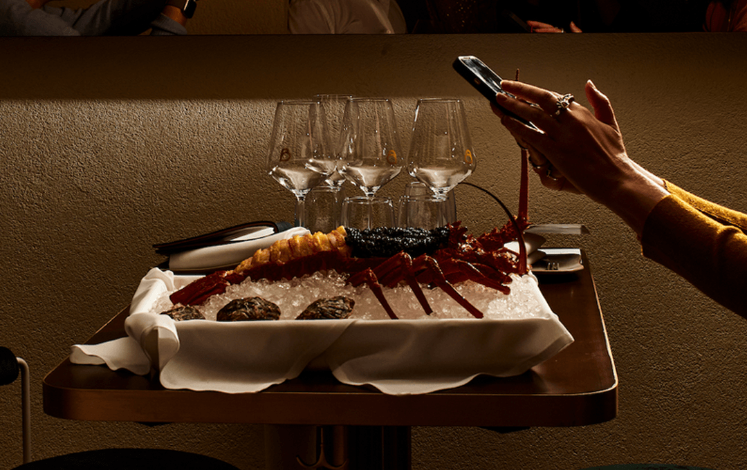 A lobster on a table with a person taking photos on their phone.
